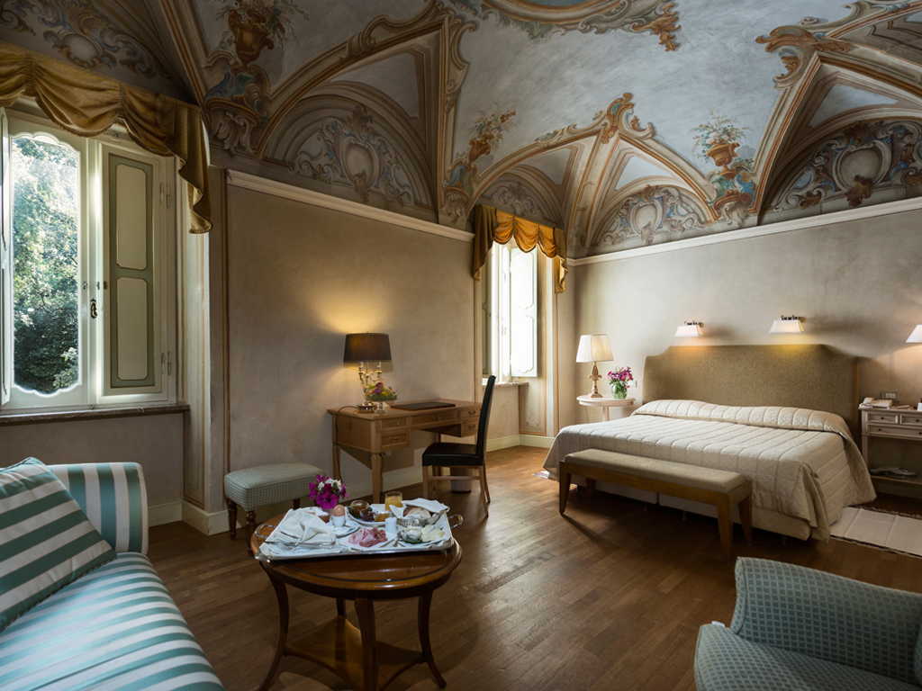 Hotel Alla Posta dei Donini. La camera da letto più prestigiosa della residenza d’epoca. Il soffitto, attribuito alla scuola di Francesco Appiani, affrescato con degli elementi architettonici, elementi floreali e tratti di cielo con angeli. La stanza in passato era la sala da pranzo dei Donini.