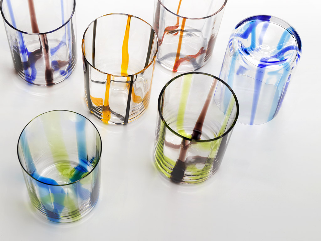 Zafferano. La linea di bicchieri “Tirache”, è arricchita da una serie di sei coppette, oggetti multifunzione, insieme ai set coordinati di bottiglie e caraffe. Dal fondo del bicchiere si inanellano linee di colori alternati, bicromie filanti che attraverso una particolarissima lavorazione segnano lo spessore e impreziosiscono le forme, fino a farne vibrare il bordo lucente, in vetro trasparente soffiato e lavorato a mano.