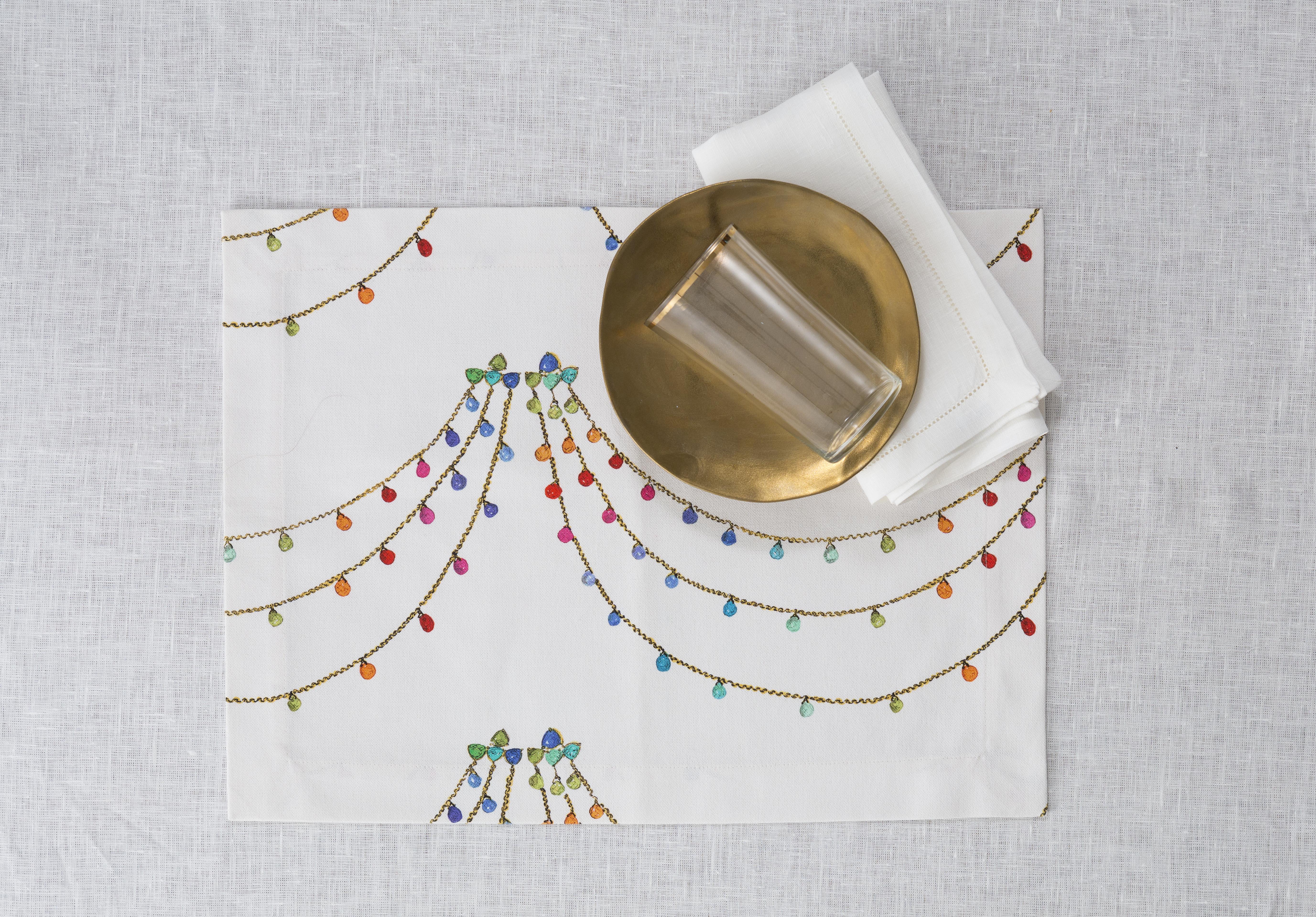 Milou Milano. Una cascata di pietre preziose e catene d’oro per “The Jewel Collection”, un decoro caratterizzato dal tratto sfumato pittorico e dai colori caramella. Disponibili tovagliette americane, tovaglie su misura e runner in puro cotone da abbinare a tovaglioli bianchi o colorati in lino con orlo à jour.