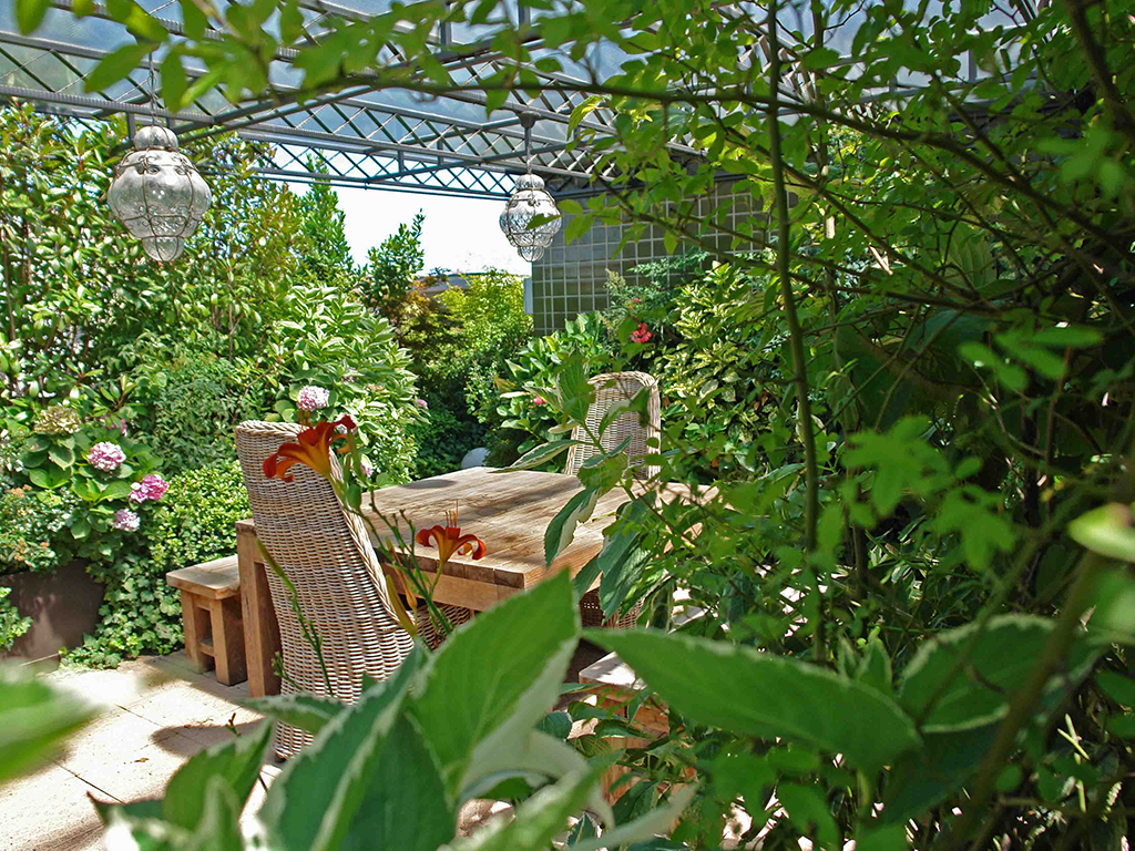 Paghera. Un terrazzo cittadino è stato completamente trasformato creando due stanze vegetali collegate fra loro da ombreggianti treillage in ferro e vetro. La prima stanza, destinata al relax e alla conversazione, è arredata con comode poltrone e divani avvolti dal verde. La seconda (nella foto) è una sala da pranzo con barbecue per cene open air. L’illuminazione d’atmosfera per questa ricca terrazza verde è affidata a lanterne di vetro soffiato e fiaccole di rame, mentre coni di luce calibrati in base all’altezza delle piante garantiscono una distribuzione più tecnica della luce.