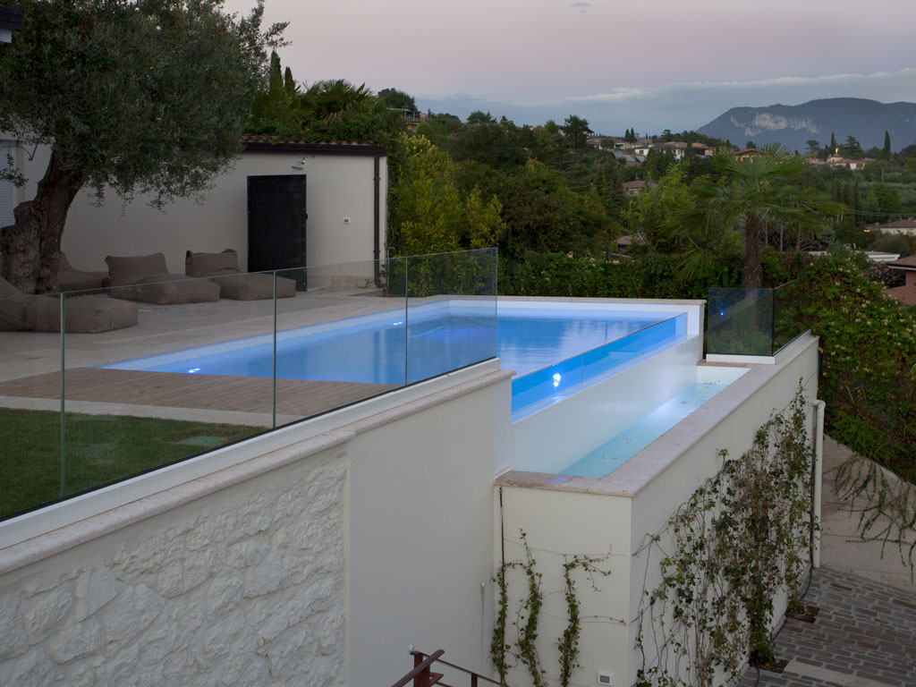Isola Verde Piscine. Due vasche con vista sul lago di Garda, una principale da 8 x 4 mt e una minipiscina idromassaggio da 2,20 x 2,20. Le due piscine fanno parte della stessa installazione ma con impiantistica differenziata. Per entrambe è stato utilizzato il vetro temperato stratificato a costituire parte della struttura. L’impianto di riscaldamento e la chiusura a tapparella della vasca principale garantiscono l’utilizzo delle piscine durante tutto l’anno. Una APP consente inoltre il controllo da remoto.