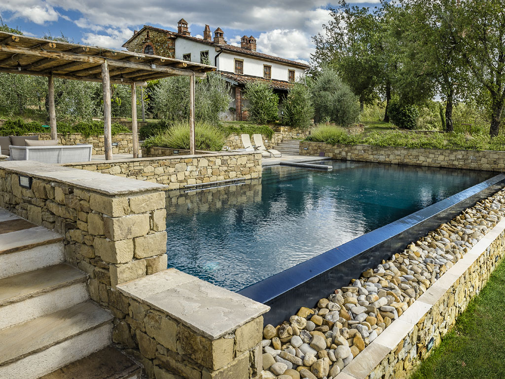 Piscine Castiglione. Circondata dalla natura toscana e da colline di ulivi, questa villa con piscina progettata dall’architetto Mario Baldini si integra perfettamente al contesto naturale. Lo specchio d’acqua - che misura 15 x 5 metri con una profondità che raggiunge i 2,40 metri sul lato provvisto di trampolino - esibisce forme geometriche declinate ad accomodare la pendenza naturale del terreno. Sfoggia infatti un bordo a cascata, una particolare soluzione di bordo a sfioro adatta per terreni con dislivello, con griglia ribassata rivestita da ciotoli in pietra locale.