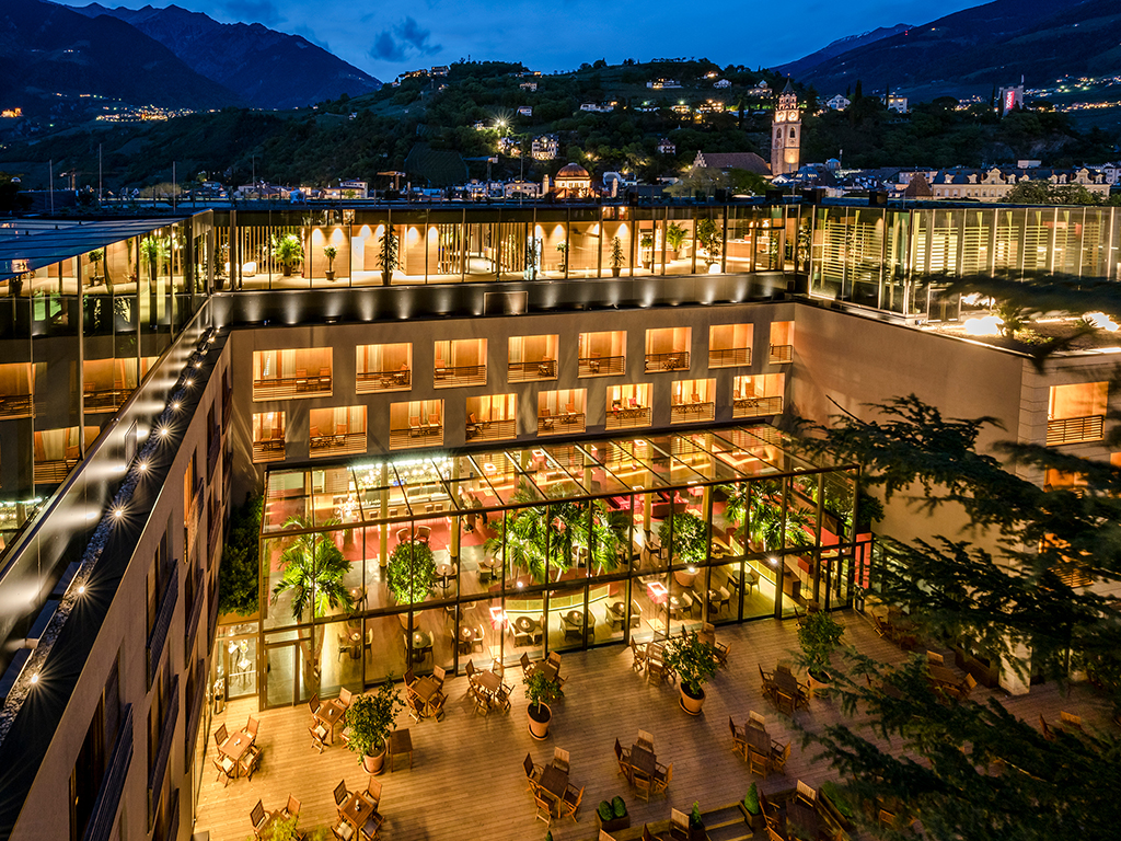 Hotel Terme Merano. Un’immagine notturna della struttura. In primo piano l’area tutta vetrate della Palm Lounge, aperta tutti i giorni dalle 17 alle 24. I cocktail sono opera dello Chef de Bar Aaron Stecher.