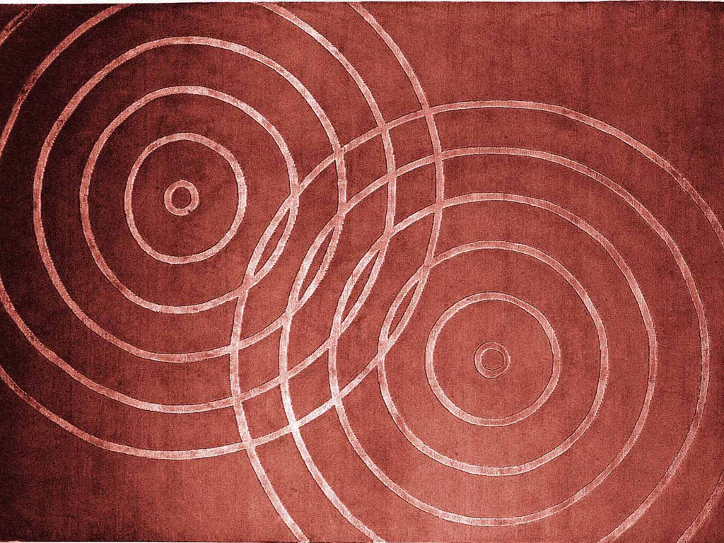 Illulian. Nell’immmagine il tappeto rosso “Drips”. Le increspature circolari, tono su tono, rompono l’apparente quiete di una superficie d’acqua. Appartenente alla linea “Design Collection” è realizzato a mano in lana himalayana, pura seta e colori vegetali. Dimensione: su misura.