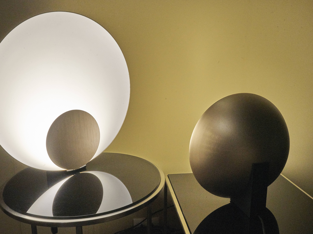 Oluce. La lampada da tavolo “Siro”, progettata da Marta Perla è composta da una lente e da un disco frontale che libera - grazie ad una sottilissima fonte LED - una luce radente al suo interno. Se vista frontalmente, “Siro” è costituita soltanto da due elementi bidimensionali, suscitando l'illusione di essere in perfetto equilibrio sul piano. In due dimensioni e due finiture: bronzo satinato oppure oro satinato, con curva interna della calotta in bianco.