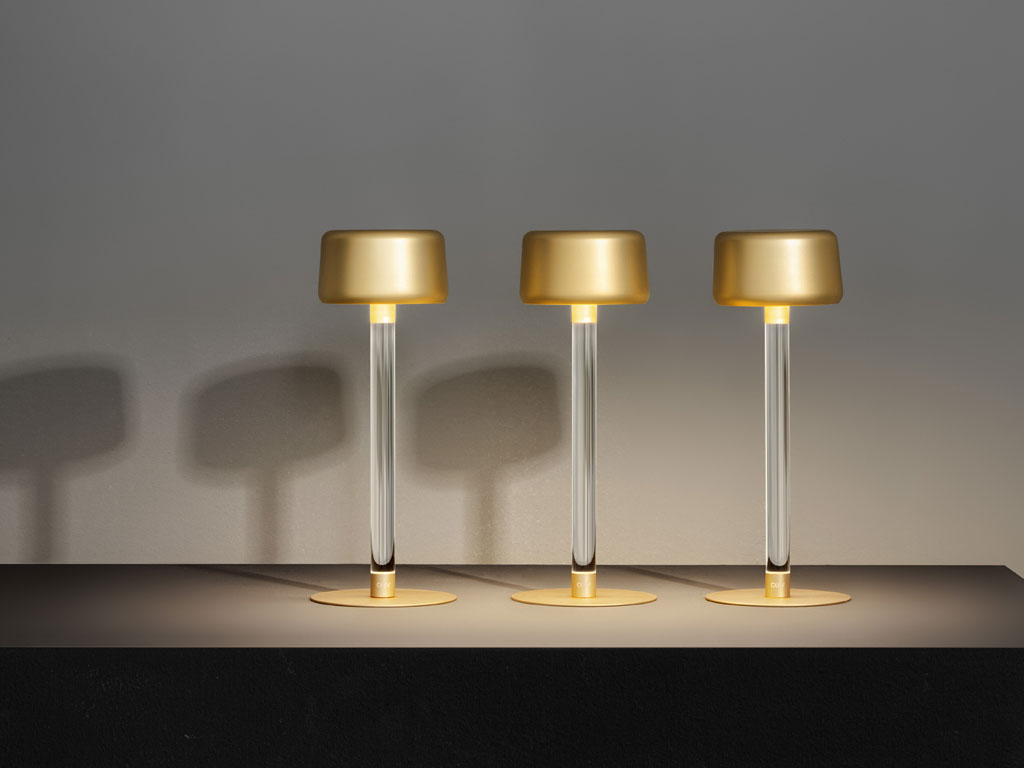Olev. “Tee”, è una lampada da tavolo senza fili, a batteria ricaricabile, Le finiture del paralume e della base circolare sono disponibili in oro ricco, nero, bianco brunito e titanio. Stelo trasparente in plexiglass. La base è magnetica, per essere posizionata a scomparsa anche sotto superfici sottili: lo stelo può così essere semplicemente appoggiato mantenendo la stabilità della lampada. La tecnologia LED Dim to Warm offre la possibilità di regolare a piacimento l'intensità e il colore della luce. Design Marc Sadler.