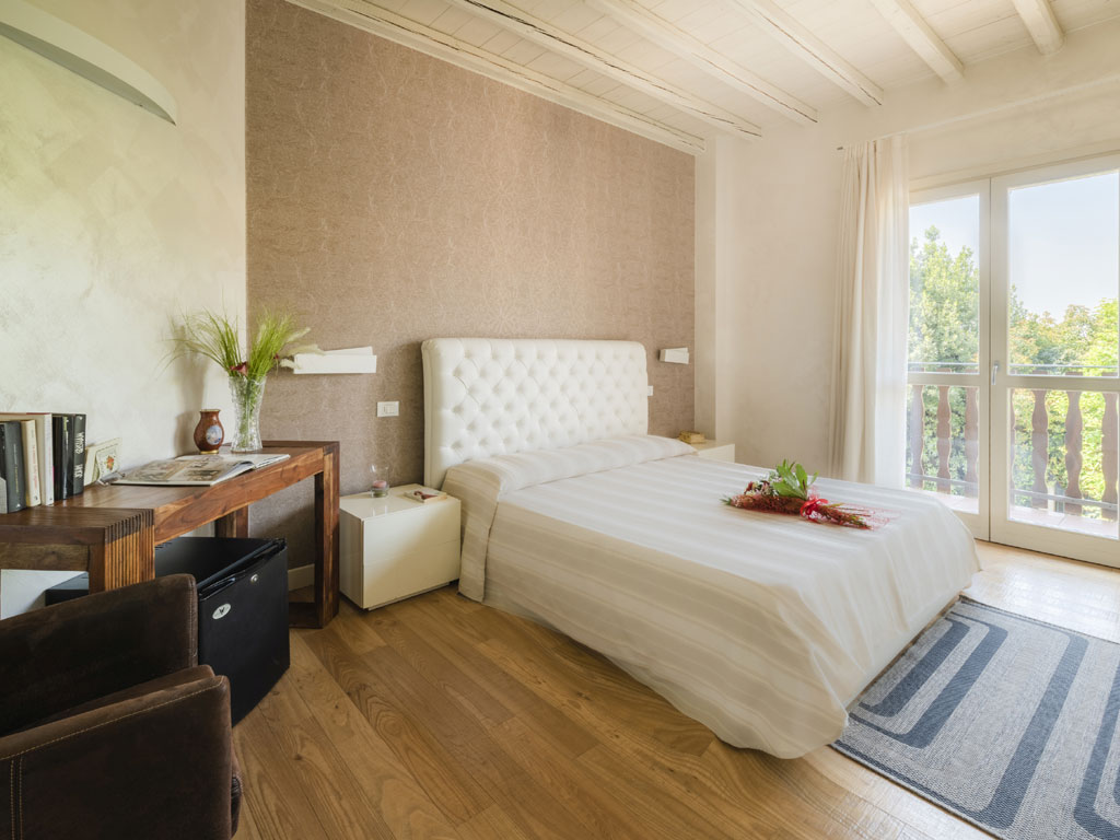Agriturismo Corte Lantieri. Camera Nike. Suite matrimoniale, dedicata alla Dea della Vittoria, composta da camera e salotto con divano/letto. Grandi dimensioni, luminosa con doppio affaccio e balconata con vista panoramica su vigneti e colline. Soffitto con grandi travi in legno e arredi eleganti e moderni.