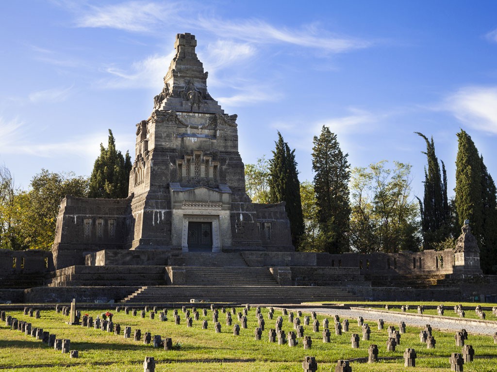 Crespi d'Adda. Il cimitero. Il cimitero ha una forma planimetrica rettangolare. L’entrata si trova a metà del lato maggiore, proprio di fronte ad un maestoso monumento funebre di forma piramidale, che ricorda in una forma rivisitata in senso industriale gli ziqqurat centro americani. Nei prati centrali del camposanto sono ordinatamente disposti i piccoli cippi, tutti rigorosamente uguali, a perenne memoria di coloro che vennero sepolti a spese dell’azienda. Le prime file sono spesso occupate da bambini.